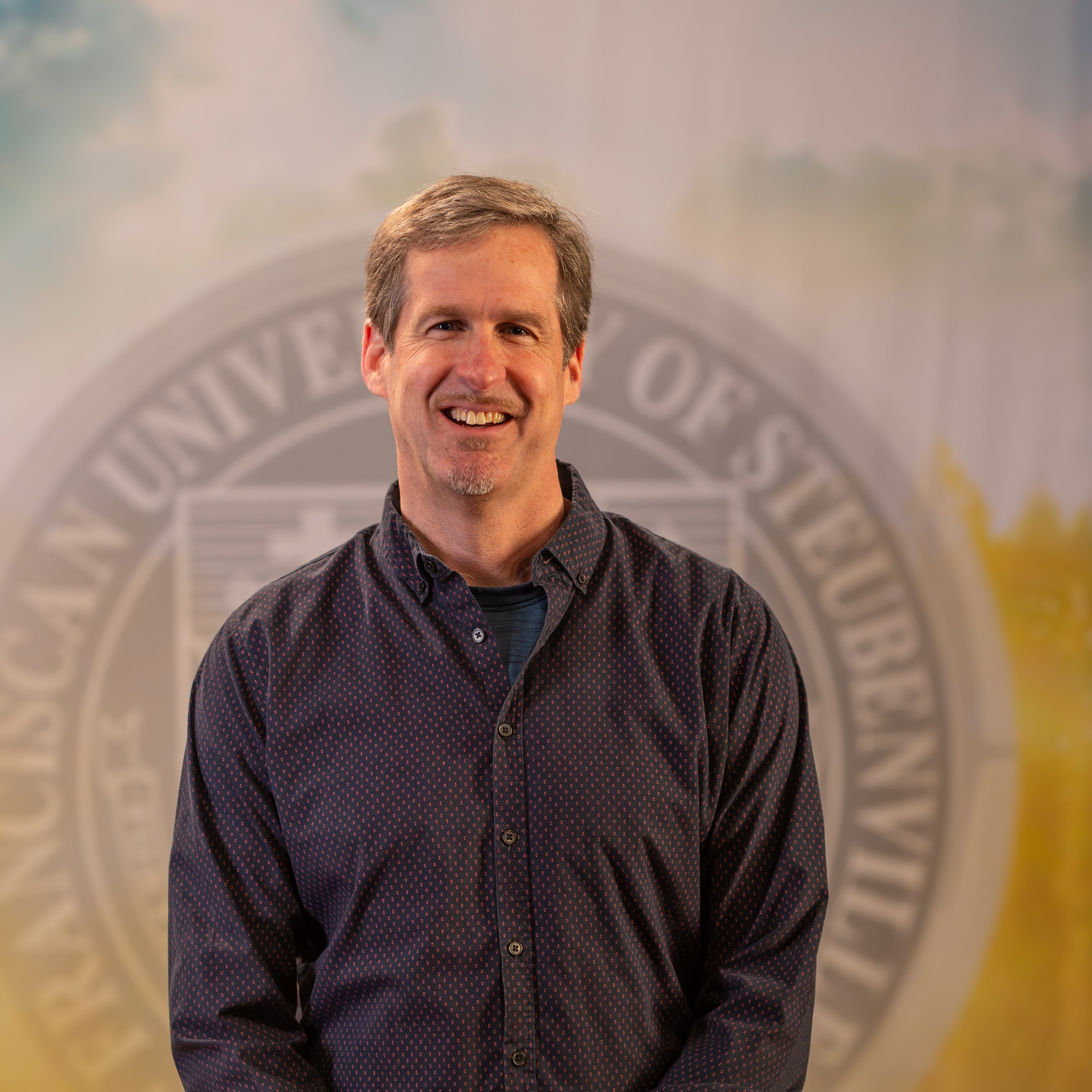 To Web – Dr. Daniel Kuebler FEB2025–2 Dr. Dan Kuebler standing in front of the Franciscan University of Steubenville seal.
