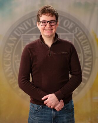 Faculty portrait of a Dr. Patrick Carzon standing in front of the Franciscan University of Steubenville seal.