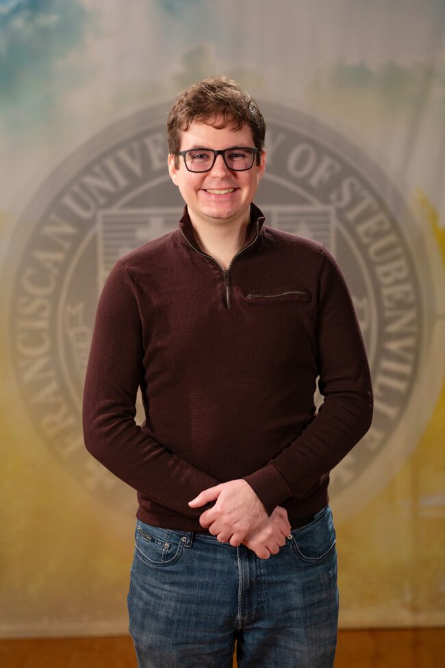 Patrick Carzon_Faculty Portrait_Spring2026_-2751 Faculty portrait of a Dr. Patrick Carzon standing in front of the Franciscan University of Steubenville seal.