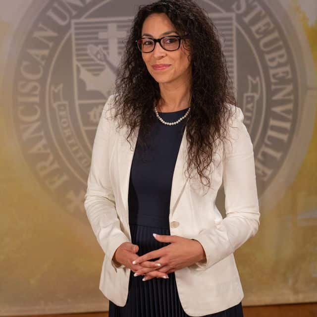 Dr Fernanda Psihas 2025 Dr. Fernanda Psihas wearing a dress and white blazer in front of the university seal.