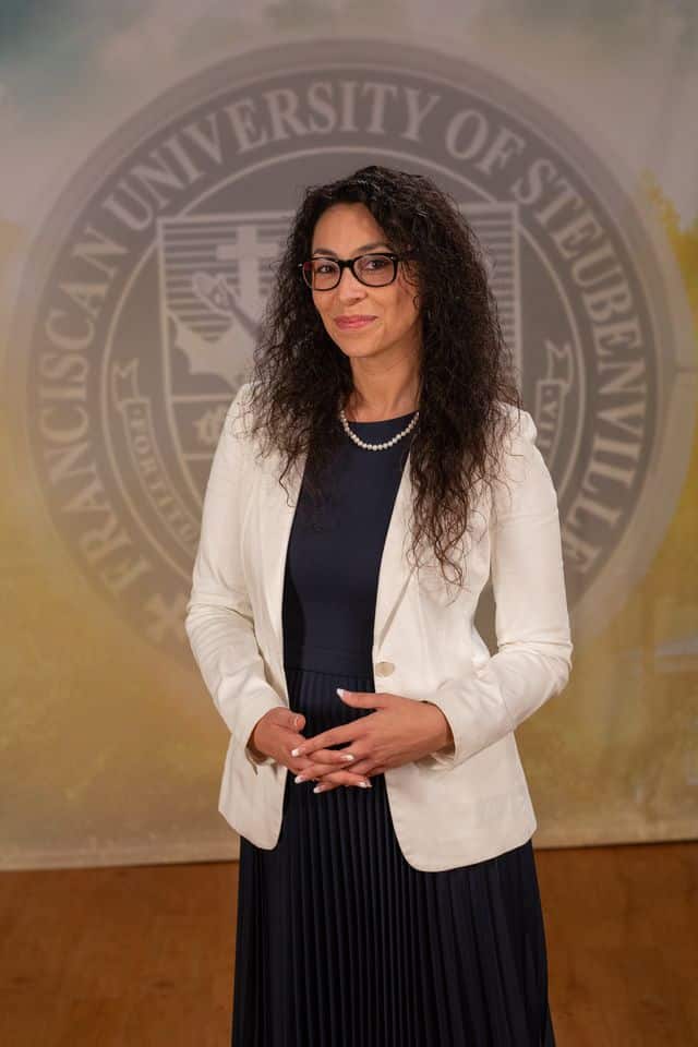 Dr. Fernanda Psihas wearing a dress and white blazer in front of the university seal.