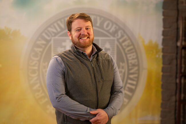 To Web – Matthew Cahill FEB2025 –3 Matthew Cahill standing in front of a college seal.