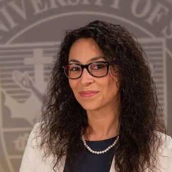 cropped-Dr-Fernanda-Psihas-2025.jpeg Dr. Fernanda Psihas wearing a dress and white blazer in front of the university seal.