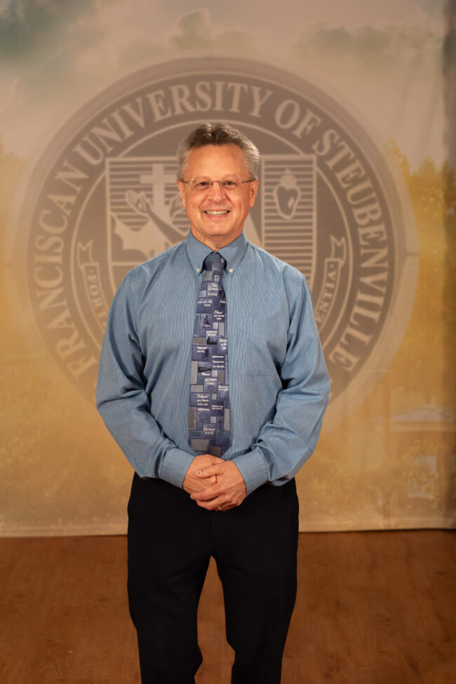 Dr. Tom Webb_FNL__B5A2265 Dr. Tom Web in a professional wear standing in front of a university seal.
