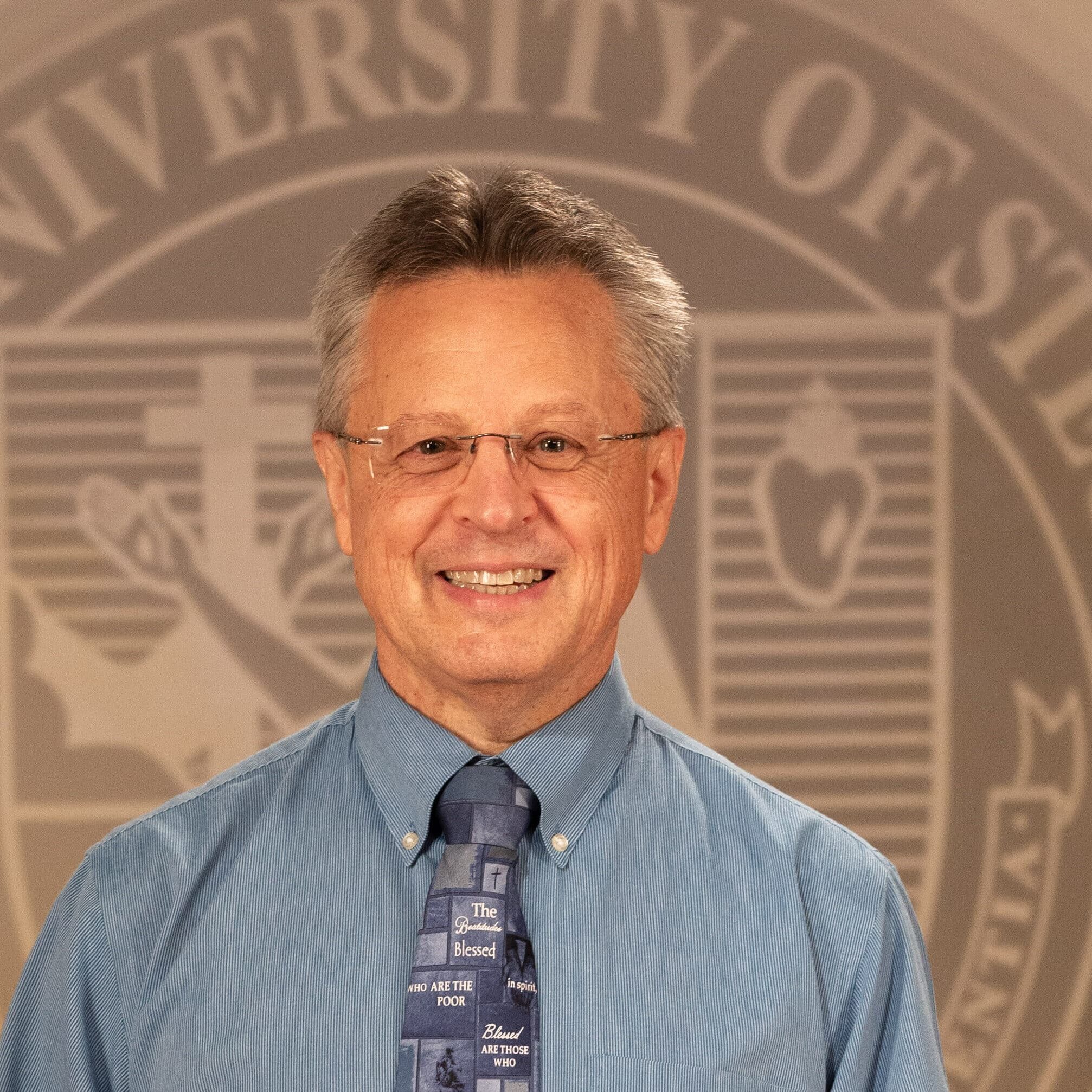 cropped-Dr.-Tom-Webb_FNL__B5A2265.jpg Dr. Tom Web in a professional wear standing in front of a university seal.